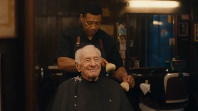 An elderly customer sits before a mirror in a warmly lit old barbershop while a dark haired barber finishes his work.