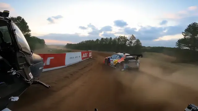 A helicopter flies low beside a speeding rally car on a dirt road, with dust trailing behind and open fields beneath a blue sky.