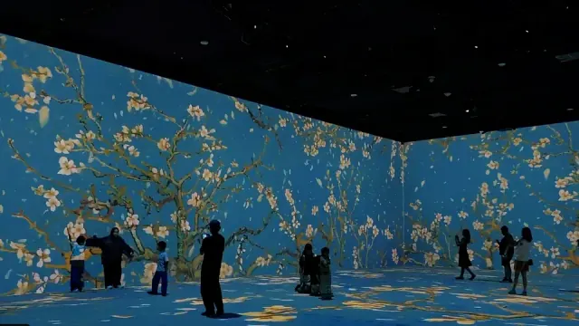 Visitors silhouetted inside a dark exhibition room covered in cobalt blue blossom projections
