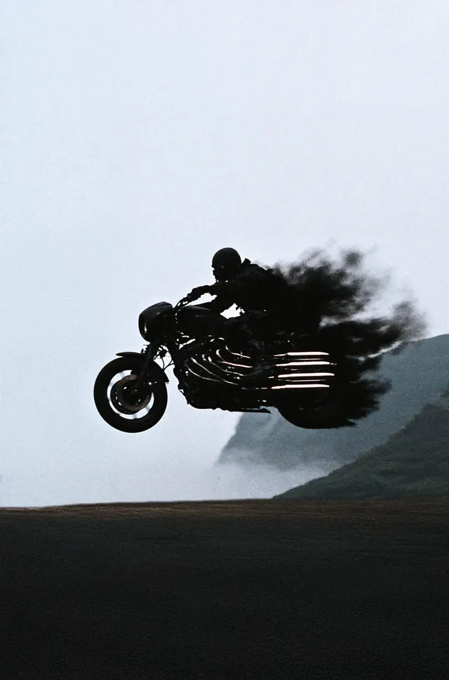 An airborne motorcycle and rider dissolving into black smoke above a misty mountain landscape.