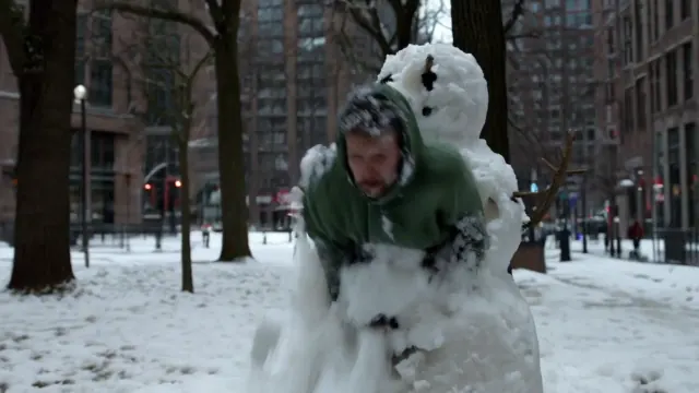 A man in a green hooded jacket emerges from a snowman on a snow covered urban sidewalk surrounded by tall buildings.