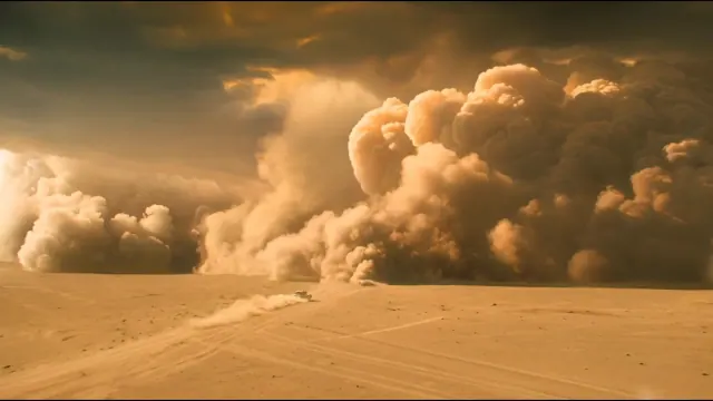 A tiny vehicle crosses golden desert sand beneath a towering wall of rolling dust that fills the horizon.