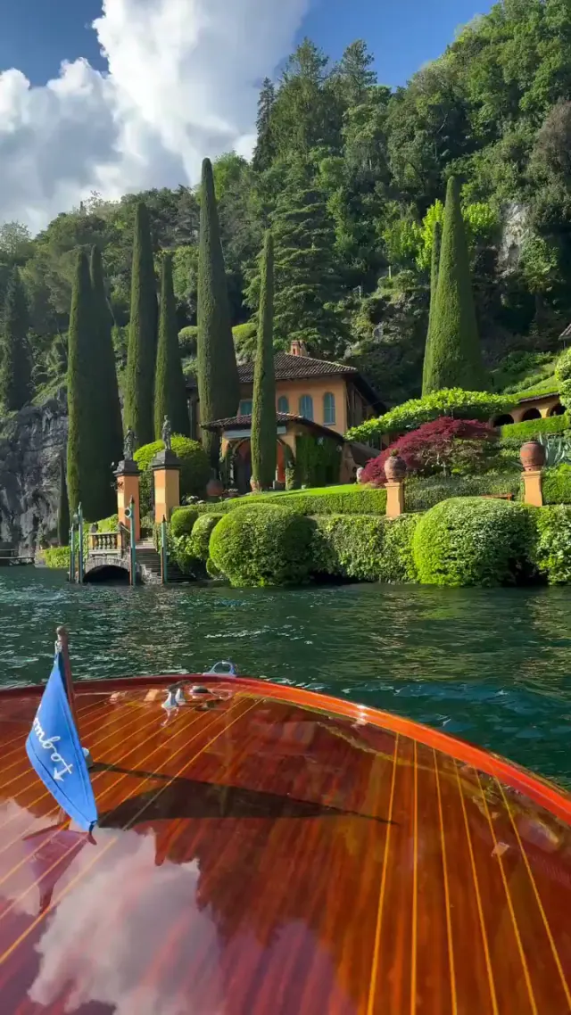 Passenger view over a polished wooden boat toward an Italian lakeside villa and garden
