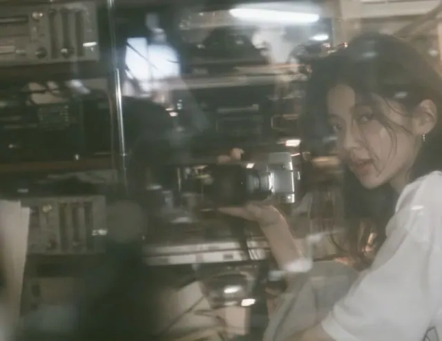 Girl in a white school shirt seated beside shelves of old audio equipment in a dim broadcasting room