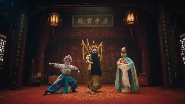 Peking opera performers in ornate gold and blue costumes stand on a warm red stage framed by dark wood and hanging lanterns.