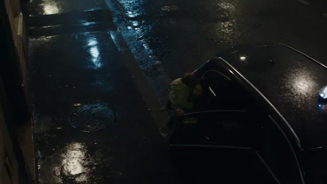 A rain soaked woman holds a small child against her shoulder on a dark city street, with wet pavement reflecting cold street lights.