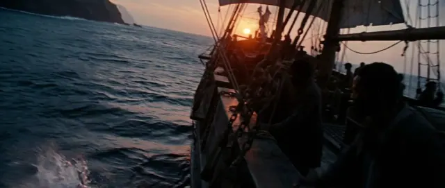 The rigging and dark hull of a large sailing ship pass close to the camera beside a rocky coast at sunset.