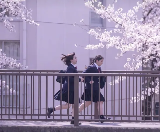 Two women in navy school uniforms running across a pedestrian bridge beneath spring cherry blossoms