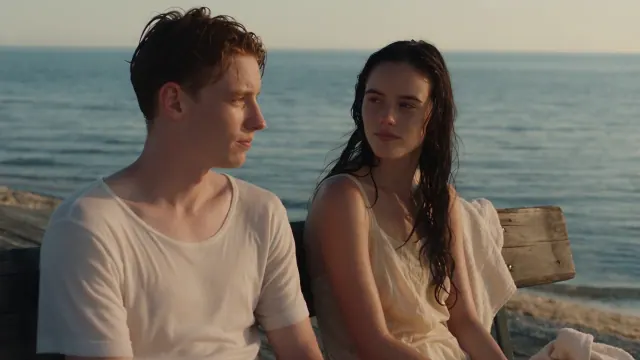 Two young adults with wet hair sit on a wooden seaside bench in warm evening light, with calm blue water behind them.
