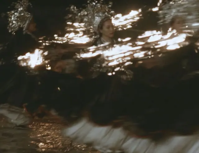 Dancer in a black pleated dress and ornate silver headdress turning through a dark nighttime scene
