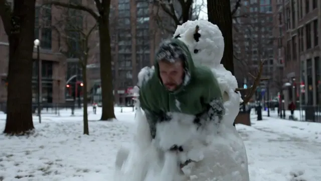 A man in a green hooded jacket emerges from a snowman on a snow covered urban sidewalk surrounded by tall buildings.