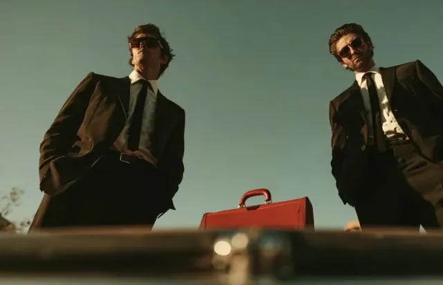 Low view from an open car trunk toward two men in black suits and sunglasses beside a red briefcase