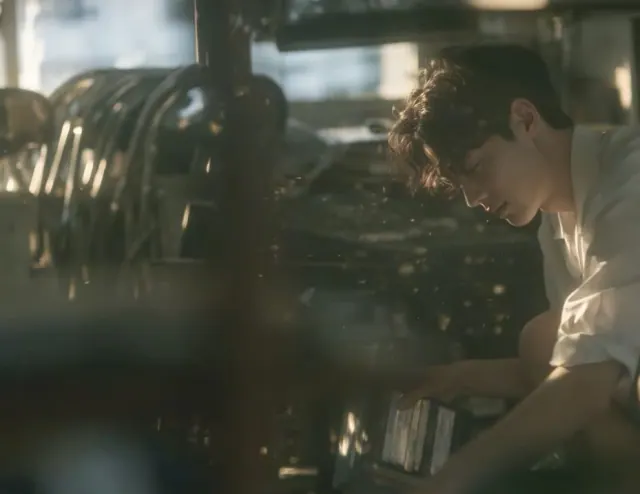 Boy in a white shirt crouching beside dusty audio equipment in an abandoned school broadcasting room